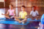 Children meditating on blue mats in a gym, wearing colorful shirts and focused expressions. Sunlight filters through large windows.