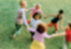 Children holding hands and spinning joyfully in a circle on green grass. Bright clothing adds color; mood is playful and lively.