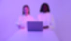 Two people in white tops stand behind a laptop on a white table. Purple lighting creates a futuristic mood. No visible text.