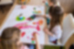 Three children painting with watercolors at a table. Bright colors on paper and palette, pencils in a cup. Creative and focused mood.