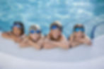 Four smiling children wearing blue goggles lean on the pool edge. Clear water glistens, creating a playful and cheerful mood.