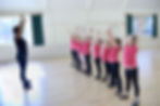 Instructor and children in pink shirts practicing dance with raised arms in a bright studio with wooden floor and green curtains.