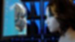Woman faces computer monitor showing a metallic robotic face. Blue and black geometric background. Icons visible on screen. Tech and curiosity.
